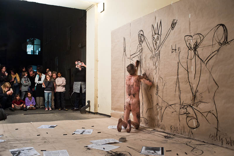 Nackter Mann zeichnet an einer Wand vor einer Gruppe Betrachter